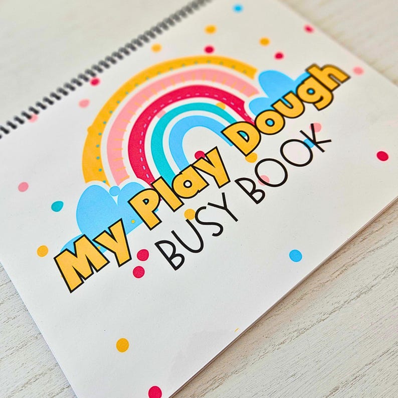 May include: A white spiral bound book with a colorful rainbow design and the text "My Play Dough Busy Book" in yellow letters.