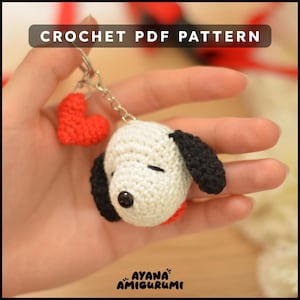 May include: Crochet PDF pattern for a white and black dog amigurumi keychain with a red heart charm. The pattern is by Ayana Amigurumi.
