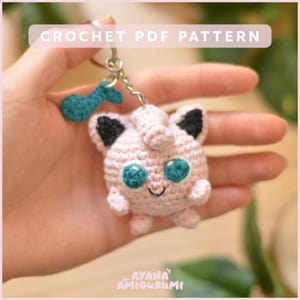 Peut inclure: Un porte-clés en forme de personnage rose crocheté avec des oreilles noires et des yeux turquoise. Le porte-clés a une chaîne argentée et un charme de note de musique turquoise. Le texte "CROCHET PDF PATTERN" est en haut et "AYANA AMIGURUMI" en bas.