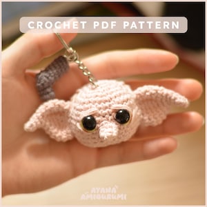 Puede incluir: Un llavero de ganchillo con una criatura rosa claro con grandes ojos negros y orejas puntiagudas. El llavero tiene una cadena plateada y un apéndice gris. El texto "CROCHET PDF PATTERN" está en la parte superior.