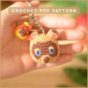 Peut inclure: Un porte-clés crocheté représentant un personnage tanuki avec des détails bruns et beiges, des yeux bleus et une petite bourse jaune avec un ruban rouge. Le porte-clés est attaché à une chaîne argentée. Le texte en haut indique "CROCHET PDF PATTERN".