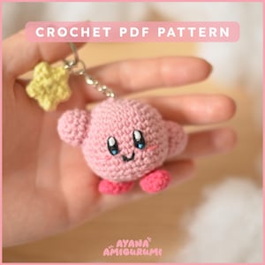Peut inclure: Porte-clés en crochet rose représentant un personnage avec un visage souriant et de grands yeux. Le porte-clés est attaché à une chaîne argentée et un charme en forme d'étoile jaune. Le texte "CROCHET PDF PATTERN" est en haut de l'image. Le texte "AYANA AMIGURUMI" est en bas de l'image.