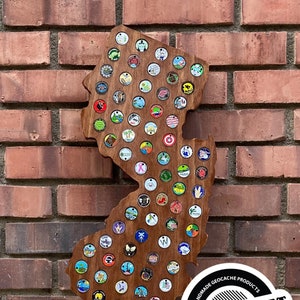 May include: A wooden outline of the state of New Jersey with a collection of colorful geocaching trackable tags attached. The wooden outline is brown and the tags are various colors and designs. The text "CACHE CRAFT" is in a black circle with white text and a white outline. The text "HANDMADE GEOCACHE PRODUCTS" is in a black circle with white text and a white outline. The text "WWW.CACHECRAFTNJ.COM" is in a white circle with black text and a black outline.