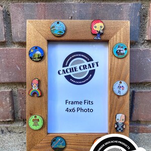 May include: A wooden picture frame with a white mat and a 4x6 inch photo opening. The frame is decorated with various colorful enamel pins, including a cartoon character, a ship, a bell, and a rainbow. The frame also features a circular sticker with the text "HANDMADE GEOCACHE PRODUCTS CACHE CRAFT WWW.CACHECRAFTNJ.COM".