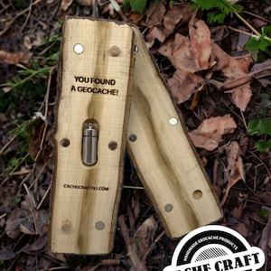 May include: A wooden geocache container with the text "YOU FOUND A GEOCACHE!". The container is made of light-colored wood and has a metal capsule inside. The Cache Craft logo is visible. The container is open, revealing the capsule.