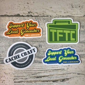 May include: Four stickers with a geocaching theme. The stickers are white with colorful designs. One sticker says "Support Your Local Geocacher" in yellow and orange letters on a blue background. Another sticker says "Support Your Local Geocacher" in yellow and orange letters on a blue background. A third sticker says "CACHE CRAFT" in black letters on a gray circle with a white background. The fourth sticker is green and says "TFTC" in yellow letters.