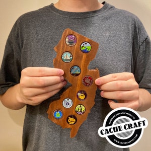 May include: A wooden outline of the state of New Jersey with 14 round, colorful, metal geocaching trackable tags attached. The wooden outline is brown and the tags are various colors, including green, blue, red, yellow, and black. The text "CACHE CRAFT" is in a black circle with a white background and the text "HANDMADE GEOCACHE PRODUCTS" is in a white circle with a black background. The text "WWW.CACHECRAFTNJ.COM" is in a white circle with a black background.