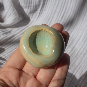 May include: A handmade ceramic ashtray with a rounded shape and a central depression. The ashtray features a glossy, light green interior and a beige exterior with a subtle, textured finish. The ashtray is held in a hand.