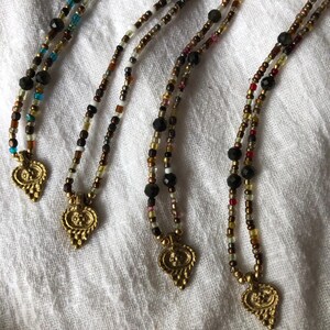 May include: Four beaded necklaces with gold pendants. The necklaces feature small beads in black, brown, gold, and turquoise. The pendants have an ornate, leaf-like design. The necklaces are displayed on a white cloth.