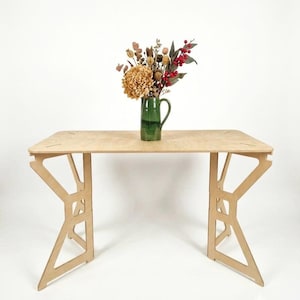 May include: A light-colored wooden desk with a flat rectangular top and two geometric, open-frame side supports. A dark green ceramic pitcher sits on the center of the desk, containing a dried floral arrangement with red berries and brown foliage.