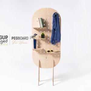 May include: A freestanding pegboard room divider with shelves and a hook. The pegboard is made of light wood and has a rounded top. The shelves are holding books, a plant, and a vase. A blue scarf is hanging from the hook. The pegboard is labelled "PEGBOARD For Home".