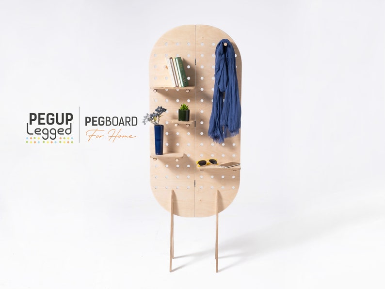 May include: A freestanding pegboard room divider with shelves and a hook. The pegboard is made of light wood and has a rounded top. The shelves are holding books, a plant, and a vase. A blue scarf is hanging from the hook. The pegboard is labelled "PEGBOARD For Home".