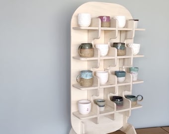 Può includere: Un'unità a scaffale in legno con una parte superiore arrotondata e un design unico. Lo scaffale è realizzato in legno chiaro e ha più ripiani sfalsati per creare un aspetto unico. Gli scaffali sono pieni di una varietà di tazze in ceramica di diversi colori e motivi.