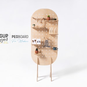 May include: A light wood pegboard with shelves and a rounded top. The pegboard is free-standing on four legs. The pegboard is decorated with small plants, a macrame wall hanging, and other decorative items. The text "PEGUP Legget PEGBOARD For Makers" is visible in the lower left corner of the image.