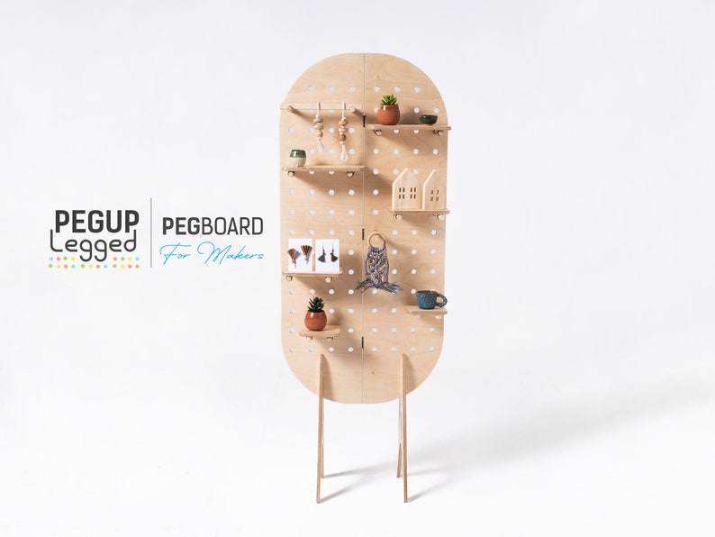 May include: A light wood pegboard with shelves and a rounded top. The pegboard is free-standing on four legs. The pegboard is decorated with small plants, a macrame wall hanging, and other decorative items. The text "PEGUP Legget PEGBOARD For Makers" is visible in the lower left corner of the image.