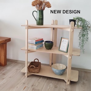 Puede incluir: Estantería de madera clara con tres niveles, que muestra libros, cuencos decorativos, una cesta tejida y una imagen enmarcada. También se ve un jarrón verde con flores secas y una pequeña planta en maceta. Se muestra el texto "NEW DESIGN".