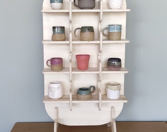 Può includere: Un'unità a scaffale in legno con una parte superiore arrotondata e tre gambe. Lo scaffale ha 12 scomparti, ognuno dei quali contiene una tazza o una tazza in ceramica diversa. Le tazze sono di vari colori e motivi, tra cui verde, blu, marrone, rosa e bianco.