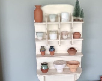 Può includere: Un'unità a scaffale in legno con più ripiani, che espone una varietà di vasi e vasi in ceramica in diversi colori e stili. L'unità a scaffale è posizionata su un tavolo in legno con due cassetti.