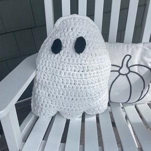 May include: White crocheted ghost pillow with black eyes. The pillow is sitting on a white wooden rocking chair.