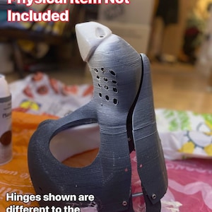 May include: A 3D printed example of a black and white mask with hinges. The text "Printed Example - Physical Item Not Included" is at the top of the image. The text "Hinges shown are different to the ones included in the download" is at the bottom of the image.