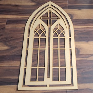 May include: A light brown wooden cathedral window cutout. The design features a cross and detailed window panes. The cutout is displayed on a dark wood surface.