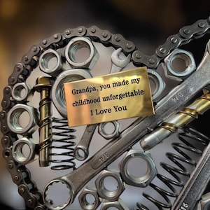 Op de afbeelding: Een gouden metalen plaquette met de inscriptie "Grandpa, you made my childhood unforgettable I Love You" omringd door een hartvormige opstelling van metalen moeren, bouten en gereedschap.