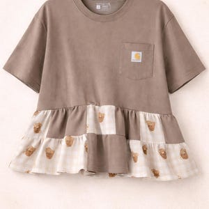 May include: A brown t-shirt with short sleeves and a pocket. The lower section has a patchwork design with bear graphics on a white and beige checked pattern.