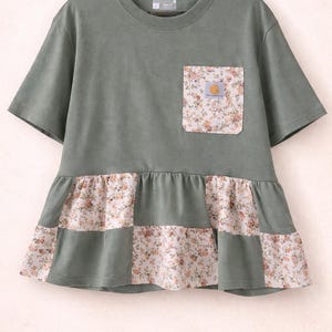 May include: Sage green t-shirt with a floral pocket and a patchwork floral and green ruffle hem. The shirt has short sleeves and a round neckline. A small logo is visible on the pocket.