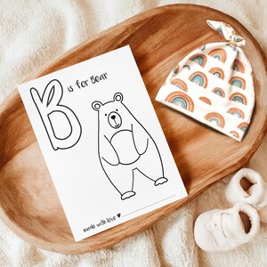 May include: A wooden tray holds a baby-themed collection. A card with a bear illustration and the text "B is for Bear" sits next to a rainbow-patterned hat and a pair of white baby booties. The items are arranged on a soft, white surface.