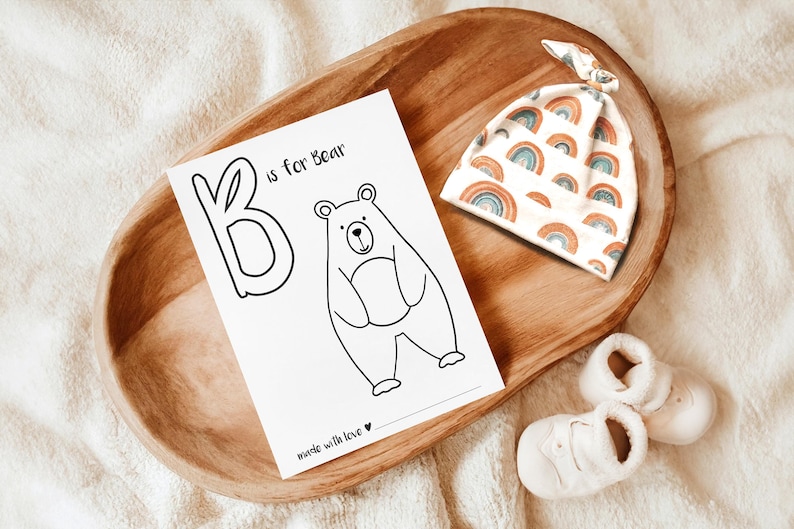 May include: A wooden tray holds a baby-themed collection. A card with a bear illustration and the text "B is for Bear" sits next to a rainbow-patterned hat and a pair of white baby booties. The items are arranged on a soft, white surface.