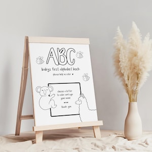 May include: A wooden easel displaying a black and white "ABC" coloring book for babies. The text includes "baby's first alphabet book" and instructions to color and sign your name. Illustrations of a koala, llama, and bees are also present.