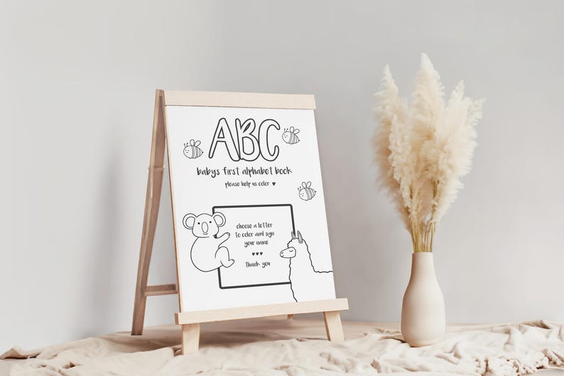 May include: A wooden easel displaying a black and white "ABC" coloring book for babies. The text includes "baby's first alphabet book" and instructions to color and sign your name. Illustrations of a koala, llama, and bees are also present.