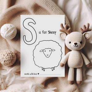 May include: A black and white illustration of a sheep with the text "S is for Sheep" on a white paper. A crocheted deer toy with brown antlers and feet is next to the paper. Two balls of yarn are also in the image.