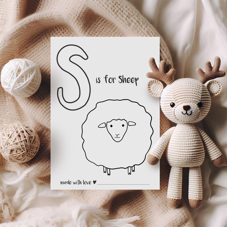 May include: A black and white illustration of a sheep with the text "S is for Sheep" on a white paper. A crocheted deer toy with brown antlers and feet is next to the paper. Two balls of yarn are also in the image.