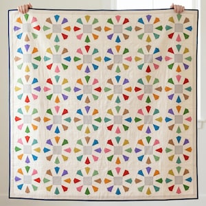 May include: A handmade quilt with a cream background and a repeating pattern of colourful geometric shapes. Each shape features a grey square centre with radiating triangles in various colours, including red, orange, green, and purple. The quilt has a navy blue trim.