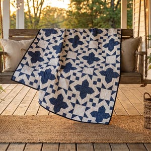 Puede incluir: Una colcha azul marino y blanca con un patrón de estrellas geométricas está sobre un columpio de porche de madera. La colcha presenta un diseño en forma de cruz azul marino sobre un fondo blanco, con un borde negro. La escena está al aire libre.