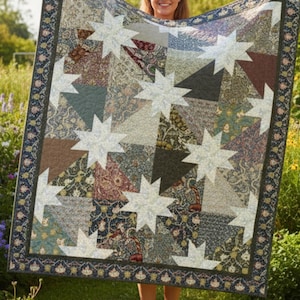 Puede incluir: Un edredón hecho a mano con un diseño de estrella. El edredón presenta un diseño de patchwork con varios patrones de tela en tonos de azul, verde, marrón y crema. El borde tiene un patrón floral. El edredón se sostiene frente a un fondo de hierba verde y flores.