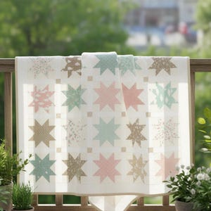 Op de afbeelding: Een witte quilt met een sterrenpatroon in roze, blauwe en bruine tinten. De quilt is over een houten reling gedrapeerd, met groen op de achtergrond. De sterren hebben bloemen- en stippenpatronen.