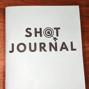 May include: A light blue journal with the text "SHOT JOURNAL" in black lettering. A black target with an arrow is above the word "SHOT".
