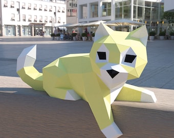 May include: A geometric paper sculpture of a cat in a resting pose. The cat is primarily yellow with white accents on the paws, tail tip, and ears. The eyes and nose are black. The sculpture is displayed outdoors on a wooden surface.