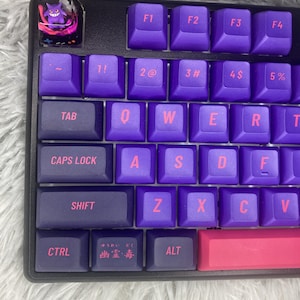 Può includere: Una tastiera meccanica viola e nera con un copritasto personalizzato che presenta un fantasma dei cartoni animati. La tastiera ha una barra spaziatrice rosa e un copritasto rosa e viola con testo giapponese.