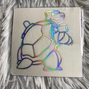 May include: A holographic sticker featuring a cartoon turtle character. The sticker has a rainbow sheen and is outlined in a dark color. The turtle is in a dynamic pose, with its arms raised. The sticker is on a square backing.