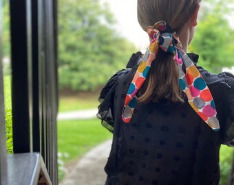 May include: A young person with long brown hair wearing a black blouse with a colorful polka dot hair tie.