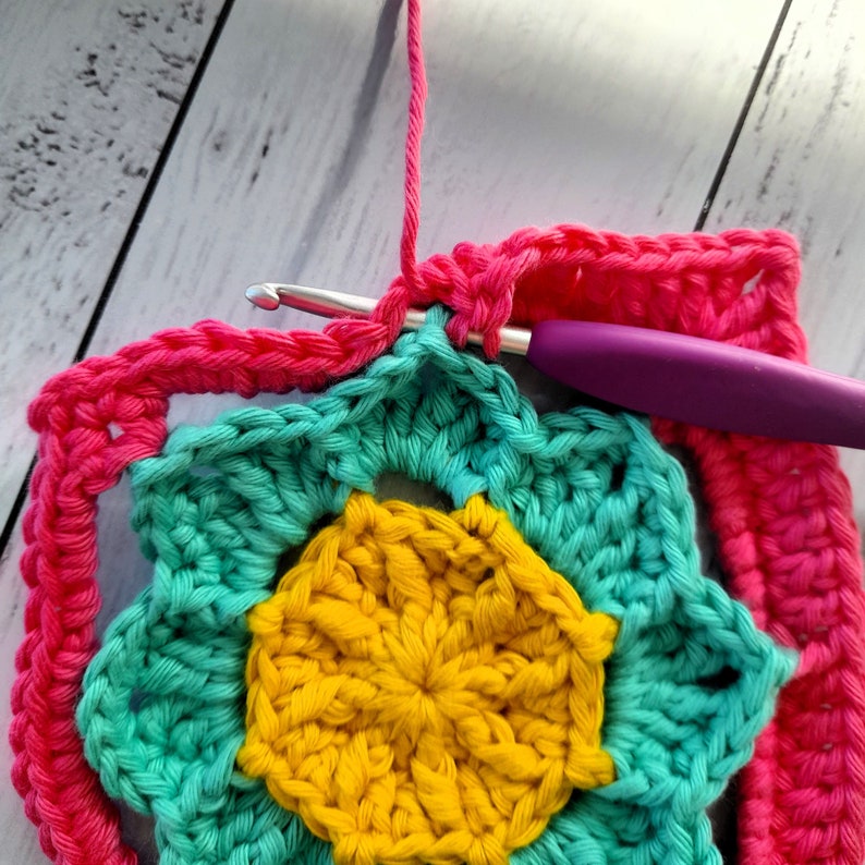 May include: A close-up of a crocheted flower with a yellow center, surrounded by teal petals and a pink border. The flower is being worked on with a purple crochet hook.
