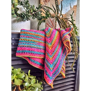 May include: A colorful crocheted shawl with stripes of pink, orange, teal, and green. The shawl is draped over a wooden surface with plants in the background.