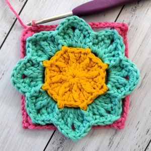 May include: A crocheted flower motif in shades of teal, yellow, and pink. The center of the flower is yellow, surrounded by a teal layer, and then a pink border.