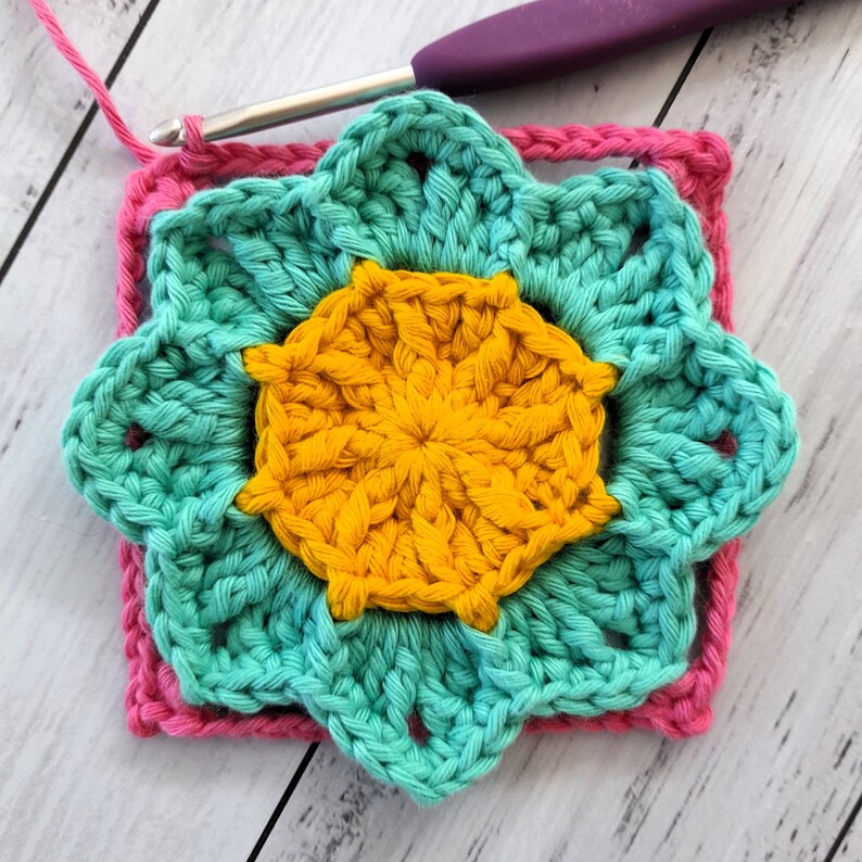 May include: A crocheted flower motif in shades of teal, yellow, and pink. The center of the flower is yellow, surrounded by a teal layer, and then a pink border.