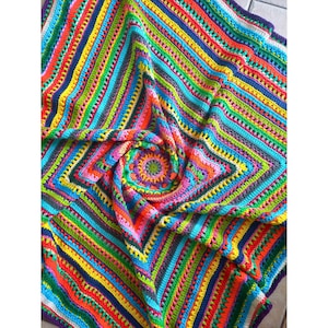 Puede incluir: Una manta de ganchillo colorida con un centro multicolor y un borde a rayas. La manta está hecha con una variedad de colores brillantes, incluyendo rojo, naranja, amarillo, verde, azul y morado.