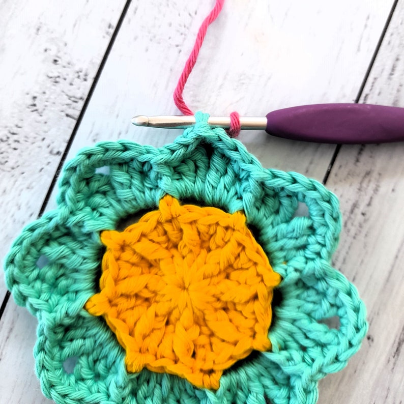 May include: A teal crochet flower with a yellow center. The flower is being worked on with a crochet hook and pink yarn.