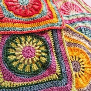 May include: Close-up of several colorful, square crochet pieces. Each piece features a central floral design in shades of pink, yellow, green, and blue, surrounded by concentric rings of various colors. The crochet work is detailed and textured.